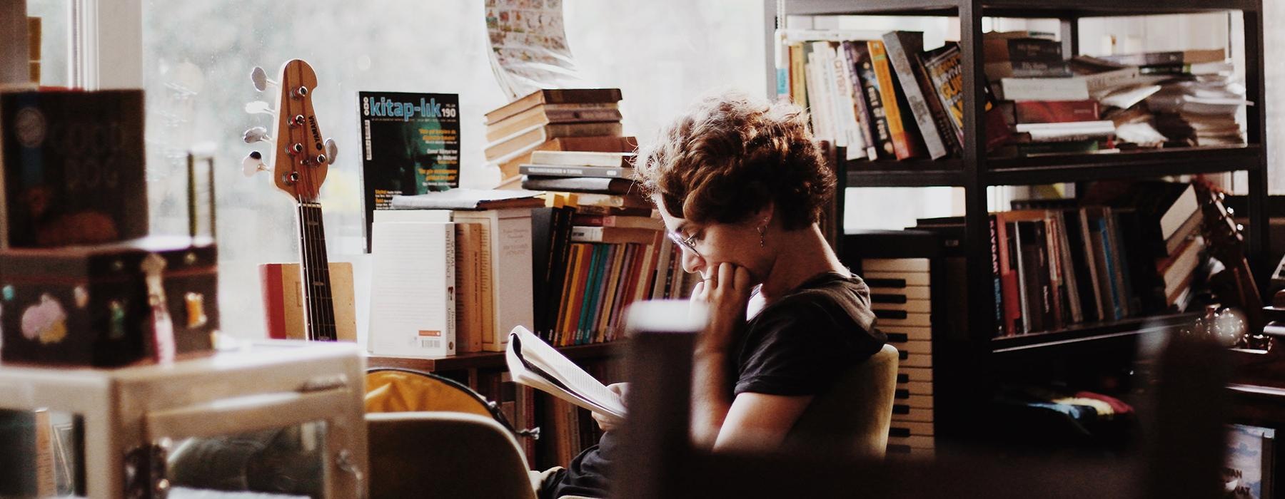 someone sitting in a chair with a book and with a bass and many books in the background